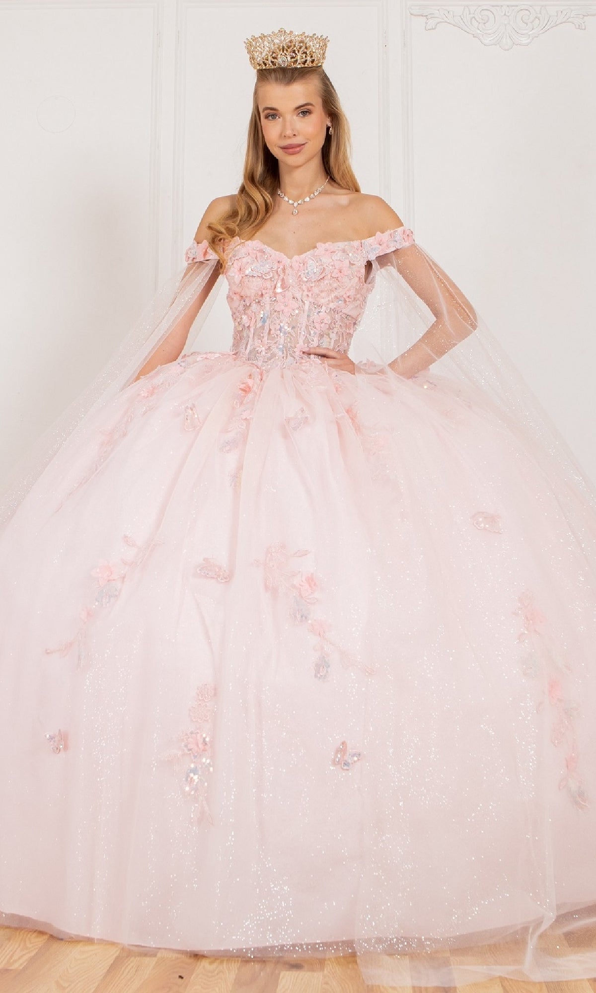 A woman in the Cinderella Couture 8075J quinceanera dress, an off-the-shoulder sparkly light pink gown with floral 3D appliques and a crown, stands against a plain white background.